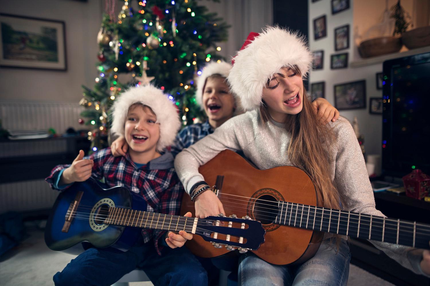 Photo of three children wearing Father Christmas hats sat in front of a Christmas tree playing guitars