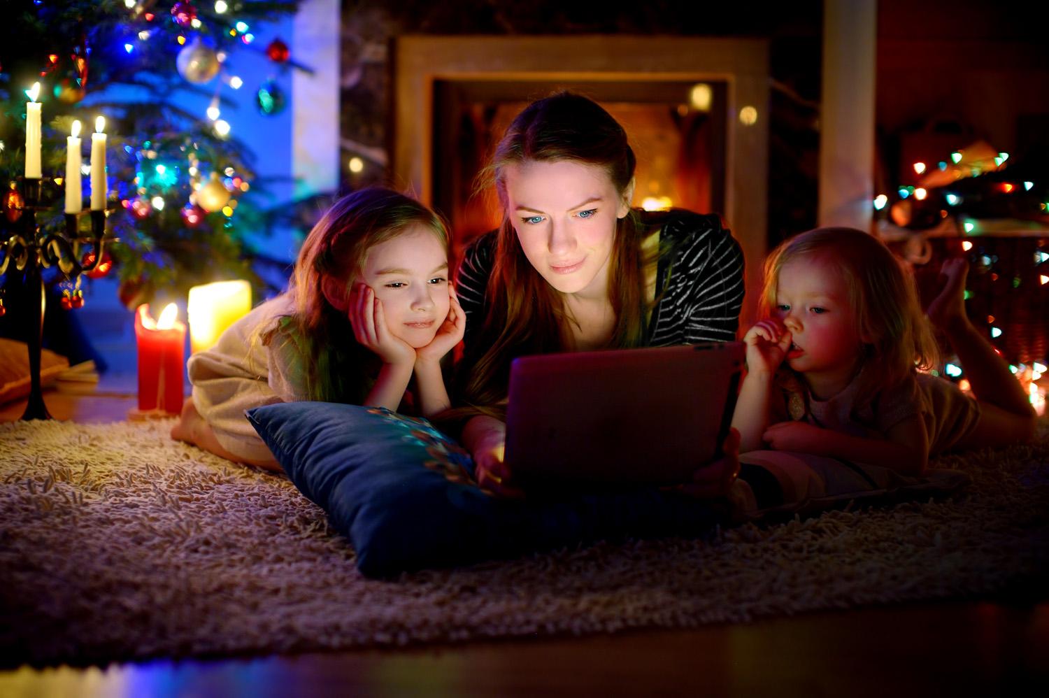 Photo of a mother and two daughters watching a Christmas film on an iPad in front of a Christmas tree